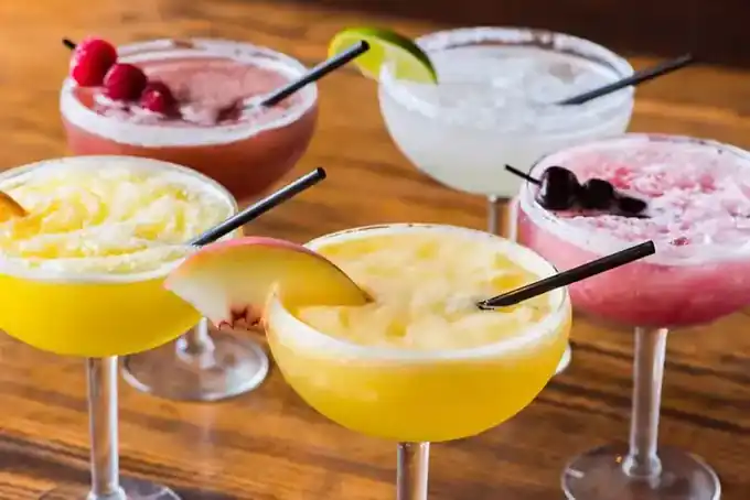 Five blended margaritas in coupe glasses, in raspberry, lime, peach, cherry and mango colors, each with its own garnish