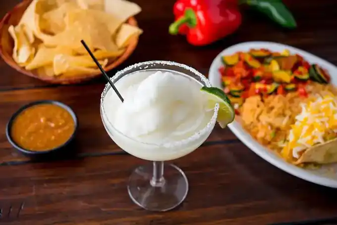 A frozen margarita in a salt-rimmed glass with a lime wedge, beside chips, salsa and a plate of rice and vegetables