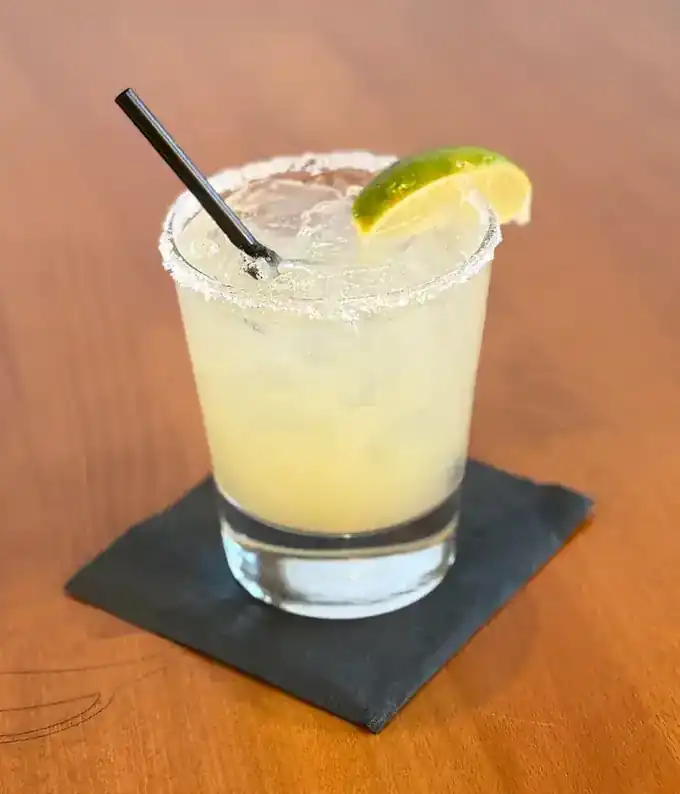 A margarita on the rocks with a salted rim and a lime wedge, set on a dark paper coaster