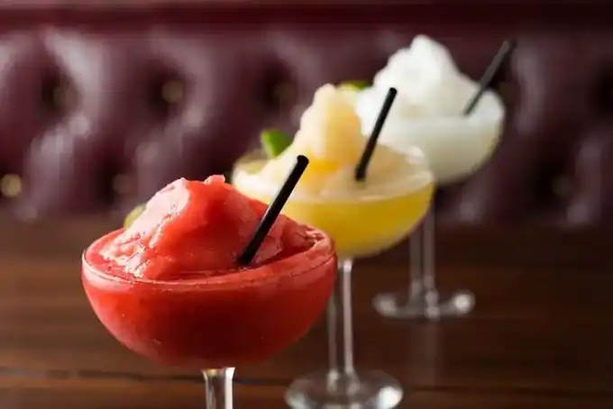 Three frozen margaritas in a row, strawberry in front, then a golden one and a white one, against a burgundy leather booth